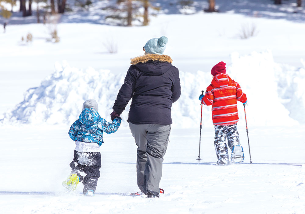 A mother snowshoes with her two children. One young child holds her hand, and the other snowshoes with poles. There are pine trees in the distance.