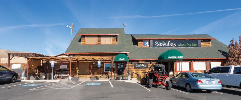 Exterior photo of Sassafras Eclectic Food Joint. Wooden building with green roof. Bar and restaurant signs on green awnings. Cars and a dune buggy type vehicle are parked in the spaces in front of the restaurant. Blue skies.
