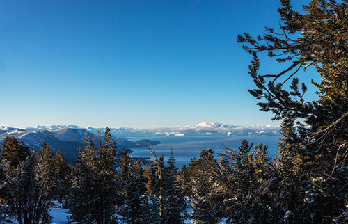 Lots of pine trees in the foreground with snow-covered mountains and Lake Tahoe in the distance. Blue skies with a few low clouds.