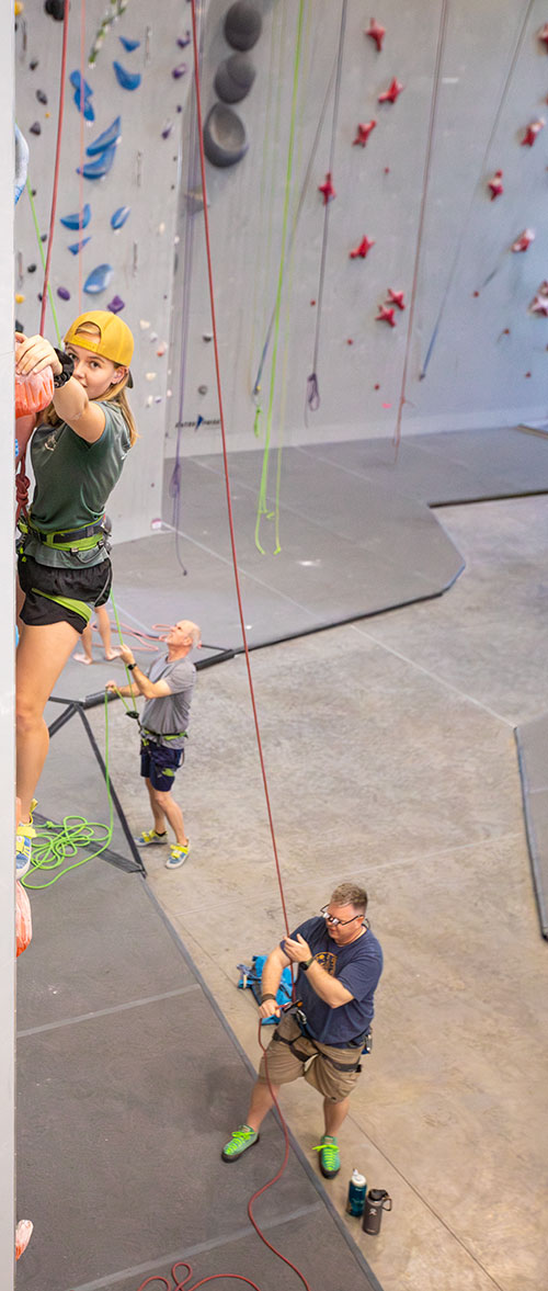 A teenage girl on the wall in a climbing gym, with an older man belaying for her.