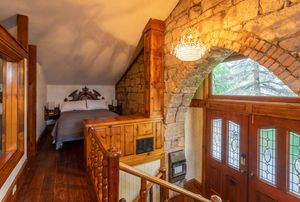 The entry to the Stone Church Lodge, from the 3rd floor. View shows stone archway over the door, a stairway leading from the doors up to the 3rd floor, where you see a large bed with ornate headboard, glass chandelier, and stairway leading down to the first floor.