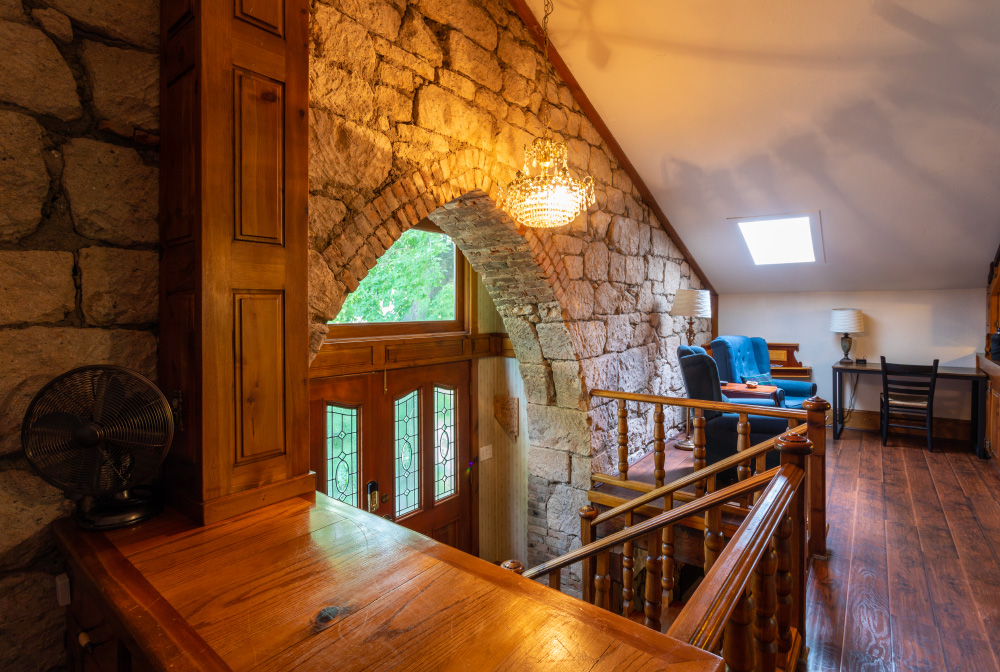 The entry to the Stone Church Lodge, from the third floor. View shows stone archway over the door, a stairway leading from the doors up to the third floor, where you see a fan, glass chandelier, two blue chairs for seating, a desk, and hardwood flooring.