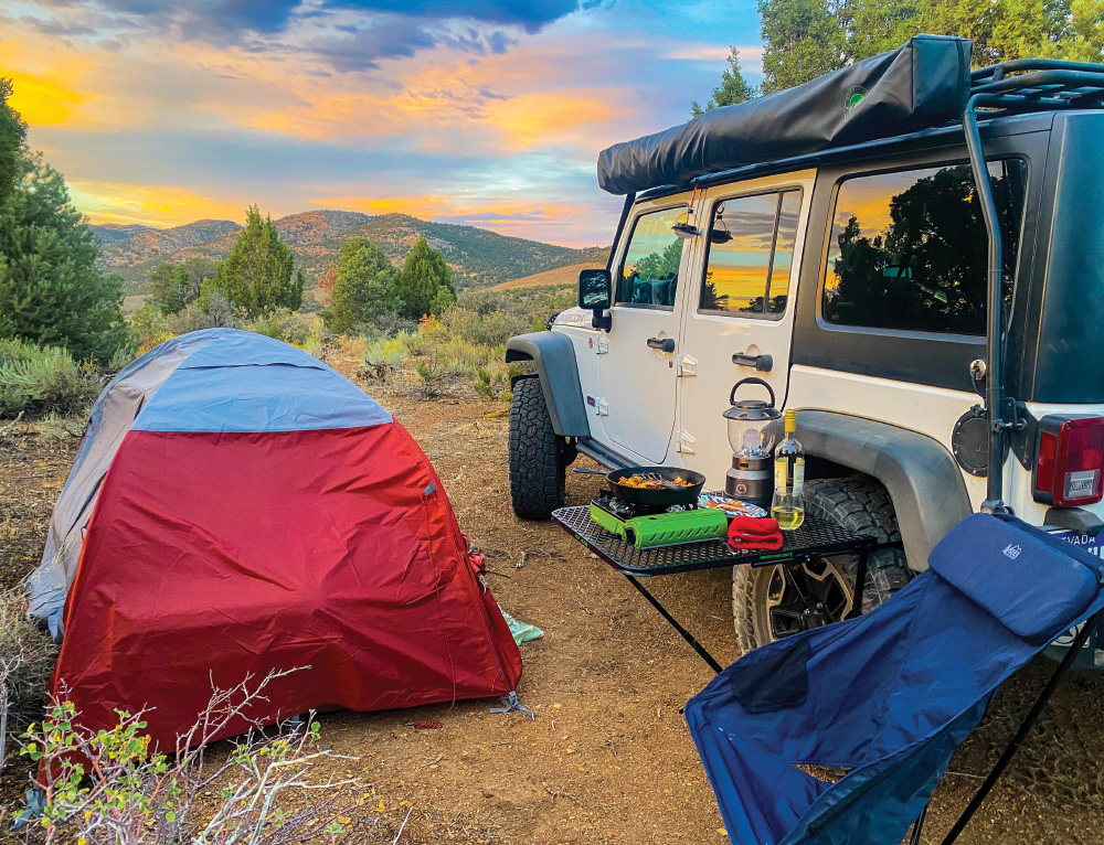A jeep is parked beside a single-person tent, in a remote part of Nevada. The tent is red and gray, the jeep is white. There is a blue camp chair sitting beside a table with some food, a glass of wine, and a lantern. The lemonade-colored sunset, yellows-orange and blue, is reflected in the jeep's windows. There are trees and sagebrush surrounding.