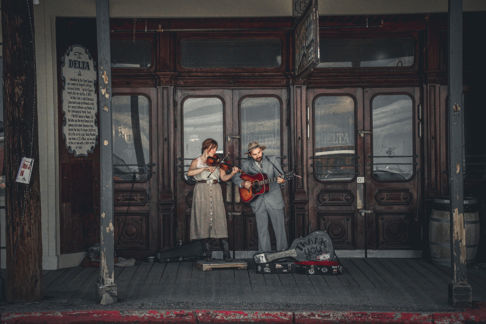 A man and a woman in period clothing play a guitar and violin. There is an open guitar case in front of them that says "thank you." They stand in front of the historic Delta Saloon in Virginia City. The sidewalks are faded gray boards and the doors to the saloon are polished dark wood and glass.