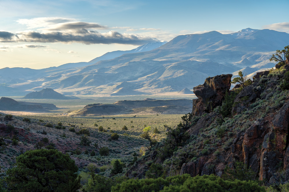 A remote valley in Nevada, lush green trees and sagebrush can be seen on many layers of mountains leading back to taller ones across the valley that still have small patches of snow on them. A thin layer of clouds is visible in an otherwise blue sky.