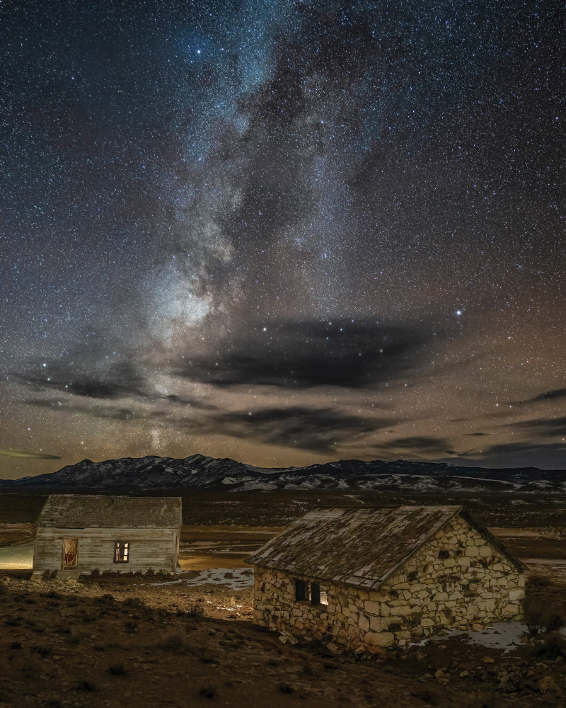 Under a starry night with a very prominent Milky Way visible, two old abandoned buildings sit in the middle of a dry valley with some snow visible on the ground. The closest building is stone and the further building is wood. There are mountains in the distance that also have snow on them.