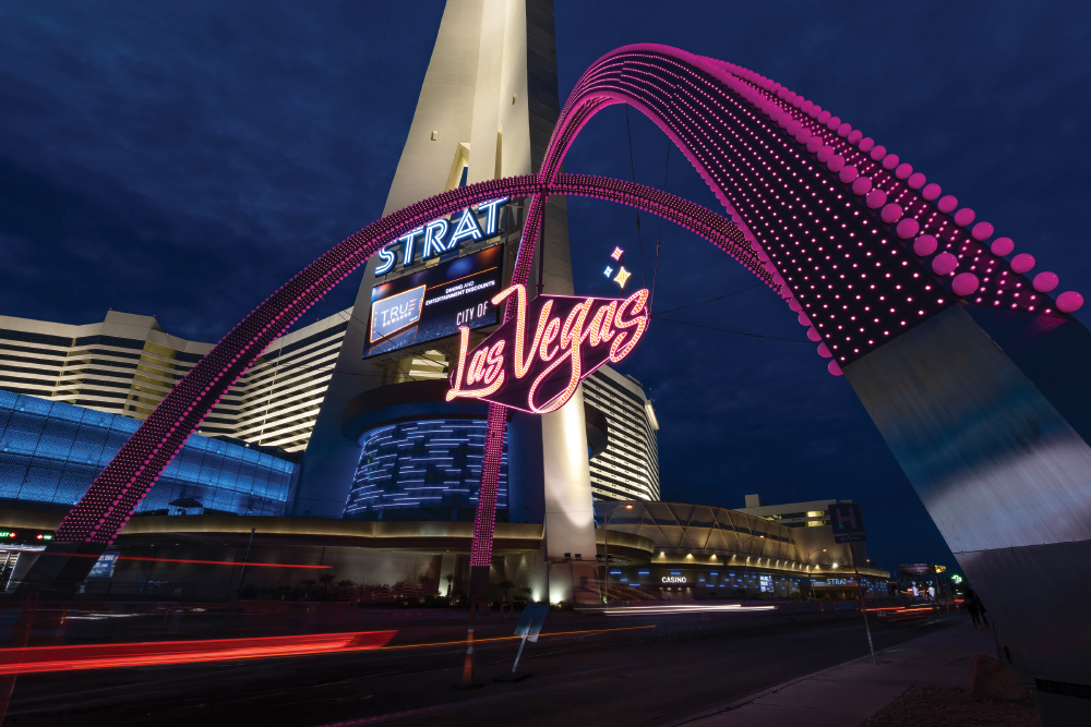 Two purple-LED-lit arches intersect over a vibrant neon Las Vegas sign. This all happens above a busy road, photographed at night with a long exposure so you can see the streaks of the brakelights and headlights. In the background is the casino The Strat, which is lit up and tall, showing hotel behind and a marquis in the foreground.