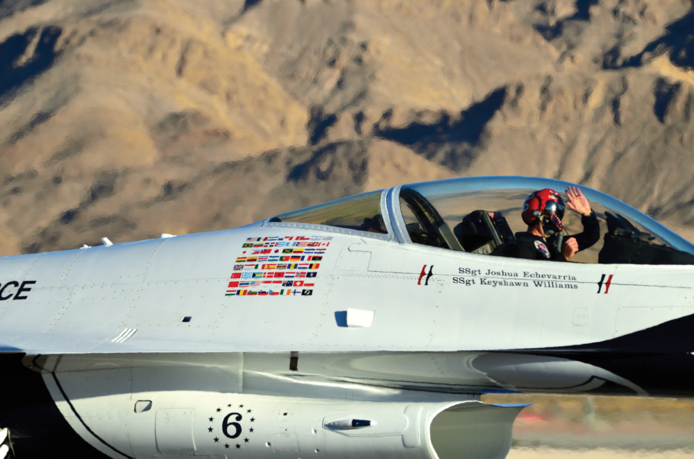 A close-up of a fighter jet in flight, with brown mountains behind. The jet pilot is waving from the cockpit, and the writing on the side reads "SSgt Joshua Echevarria" and "SSgt Keyshawn Williams"