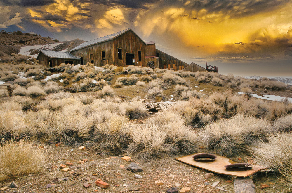 Some old junk is strewn about in the foreground, with a lot of sagebrush and some snow visible. In the distance is a large abandoned wooden building, and further out more mountains. The sky is filled with a sunset orange glow on dramatic clouds.
