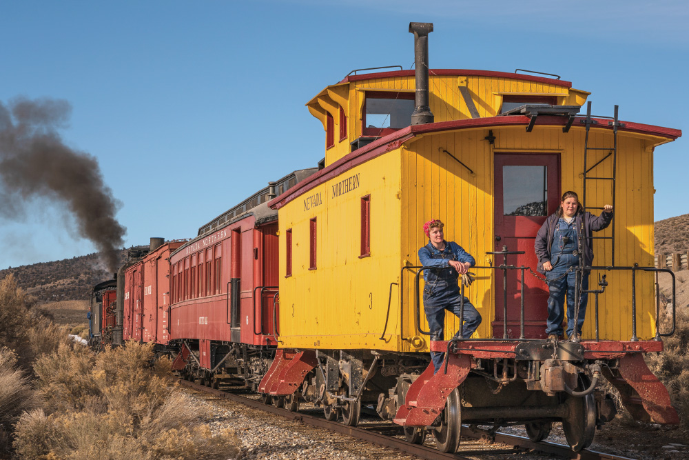 Two women, in denim overalls, stand on the back of a bright yellow caboose of a Nevada Northern Railway train. There is dark coal smoke coming from the front of the train, the rest of which is red. There is sagebrush all around, right up to the railroad tracks.