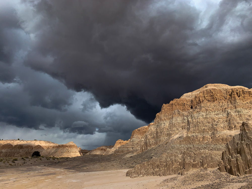 Cathedral Gorge in a storm. Tall sandstone cliffs and formations are in the foreground, tan and brown. The sky is very angry looking with dark storm clouds and rain in the distance.