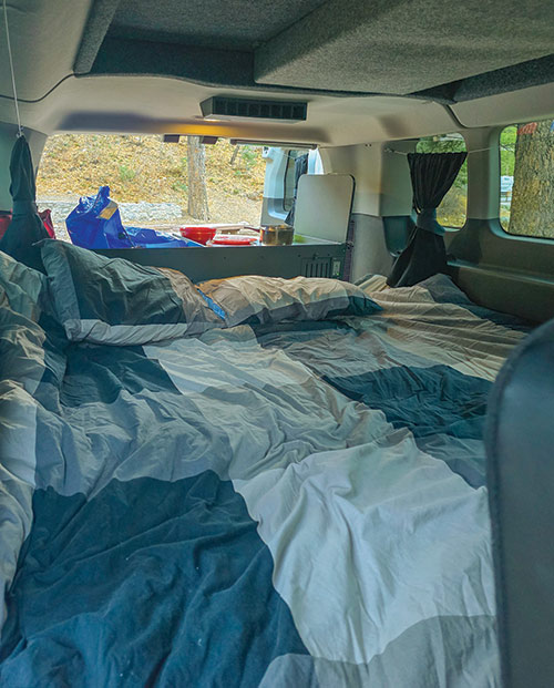 Looking out the back of a camper van, from inside. The bedding is in the foreground, and the cooking/storage area is at the back of the van.