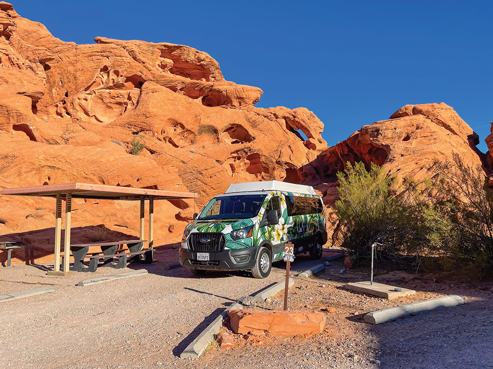 A colorful camper van sits in a campsite among tall red rocks.