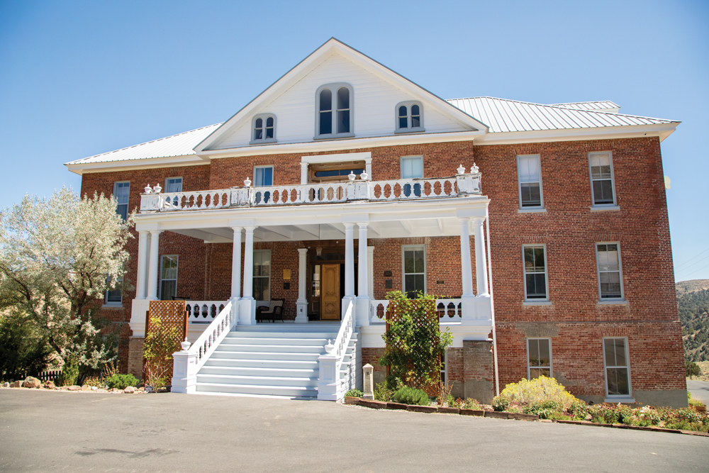 Exterior picture of St. Mary's Art Center. The front of the building has a white porch and balcony sticking out from the front which features intricate wooden rails painted white with 4 column sets with two columns in each set. The building is 4 stories tall with 16 windows on the front, and three large windows in the attic space. There are trees and other foliage surrounding the building. 