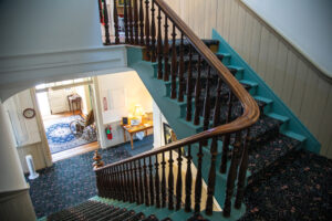 A downward shot of a staircase showing the split between the two levels. On the left, the stairs head down, and on the right, the stairs head up. The stairs are blue with a dark floral pattern carpet attached to them. The handrail is wooden with ornate spindles with a smooth, shiny finish. Down on the first floor leads to a hallway and an entry room. The carpet on the floor down below is the same carpet attached to the stairs. There is a wooden table next to the entry way with pictures and a lamp on top of it. There is an electrical box, a fire extinguisher, a sign pointing to said extinguisher above it, and a clock on the wall. The walls feature a wooden wainscot chair rail throughout both floors. The doorway to the entry is large and has three window panes on top of it. In the entry room, the floors are wooden with a blue floral rug on top of it. On top of the rug, there is an old wheelchair. On the farthest wall of the entry way sits the front door with windows next to it, and an entry table sits to the right of the door. The second story is mostly cut off in the picture, but leads to a hallway with rooms.