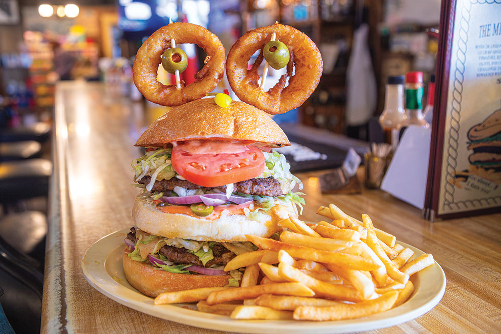 Monster burger on a plate on a countertop: Two hamburgers stacked atop one another with a third bun in the middle, with tomato, pickle, and onion. Top bun has a pepperoncini for a nose, and two onion rings with toothpics stuck through them and olives in the middle for eyes. Plate also has a pile of french fries.