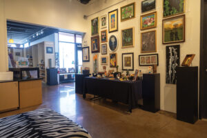 Interior of Mesquite Fine Arts Center. A gallery wall in the front right hand of the photo which showcases various types of art. There are 20 mixed media pictures on the wall. Below the pictures is a table with other various types of art pieces. In the front bottom right is an animal print seat. On the left center, there is a separate room with shelves and other art pieces adorning them. To the right of the shelves are large windows and the entrance to the art center. 