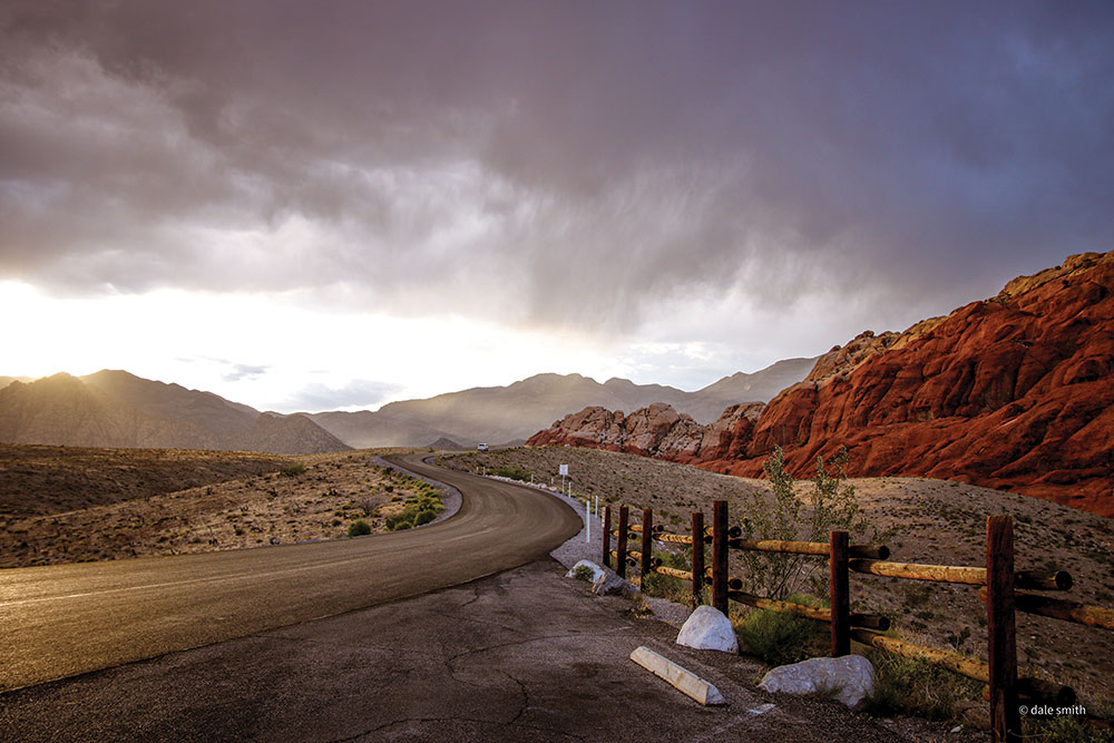 Photo taken from a pullout/parking area, a winding road leads into Red Rock Canyon, with stormy clouds in the sky.