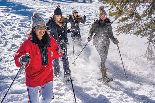 A group of four women laugh as they snowshoe with a pine tree to their right.
