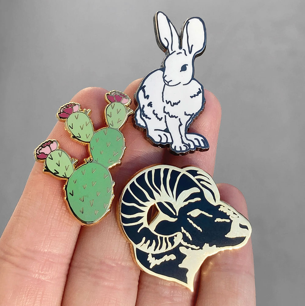 Battle Born Pins in Las Vegas, shows a hand holding three pins: one is a green cactus with red and pink blooms, another is a bighorn sheep head, and the last is a desert jackrabbit.