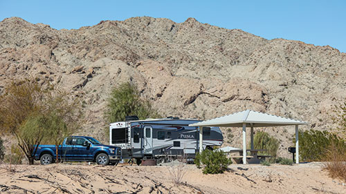 A campsite at Big Bend of the Colorado State Recreation Area, with a truck, camper, and covered picnic table and grill area. There are some trees and bushes around, and a mountain directly behind.