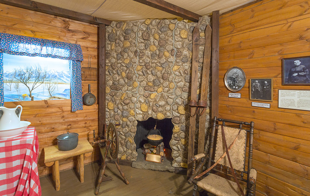 Recreation shows a fireplace with a pot inside suspended over burning logs. Wooden skis lean against the stone hearth. Photos and cooing utensils hang on the wood wall, and there is a chair, loom, small table with a pot, and larger table with a basin and red and white checkered tablecloth. The scene outside the window shows snowy winter mountains.