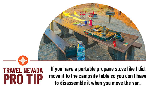 A picnic table covered with propane stove, food, and drink. Two red camping chairs are in the background. Travel Nevada PRO TIP: If you have a portable propane stove like I did, move it to the campsite table so you don’t have to disassemble it when you move the van. 