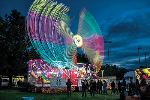 A long-exposure evening shot of a carnival ride at Carson Valley Days, with blurred lights, and people standing around in front. Photo ©Jay Aldrich