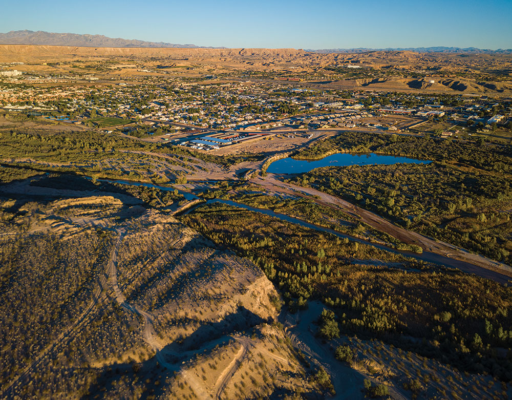 A birds-eye (drone) view of the city of Mesquite shows the Virgin River, flat-topped mesas, and a sprawling desert metropolis down below, with plenty of trees and sagebrush surrounding.