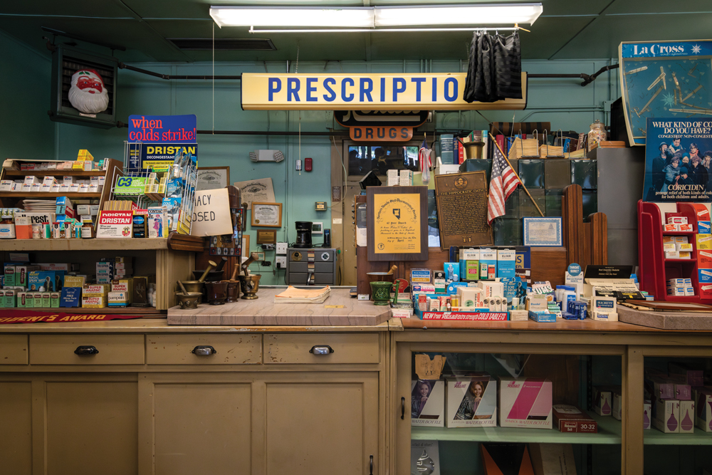 A counter full of prescription medicine sitting on top of it. Rexall's name adorns various pieces of display items for advertising purposes. A large sign sits behind the counter that says 'Prescriptions' with the 's' being covered by a hanging cloth from the light in front of the sign.