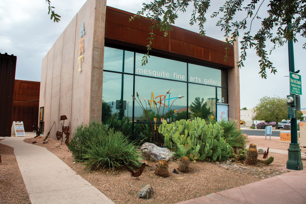 Exterior of Mesquite Fine Arts Gallery. A stone square building with the words 'ART' adorn the left side of the building. In the front of the building, there are large glass windows that read 'mesquite fine arts gallery' and the words 'ART' down below. In front of the building are various different cacti and sculptures. There is a side walk leading all around the building. The top right of the photo has leaves hanging down from a tree that isn't pictured in the photo. There is a light post in front of the building that has a sign on it that says 'Fine Arts Center. Parking in rear. Entrance off of N. Yucca St. "