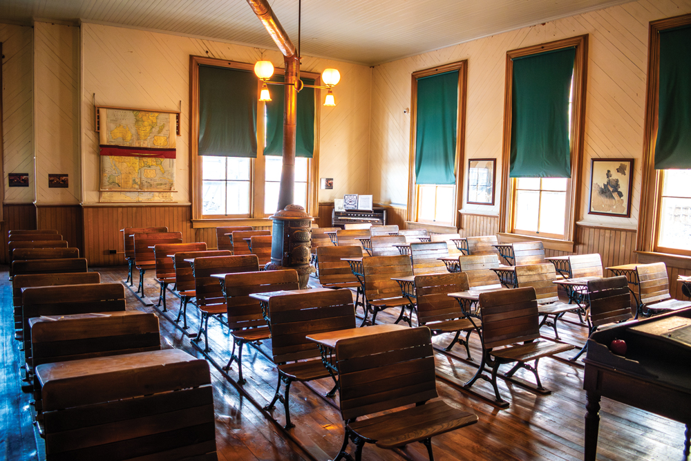 Classroom setting with rows of old wooden chair and desks lined up. A heater sits in the middle of the desks with piping that leads to the ceiling, and in front of it hangs a light fixture that holds two separate lights. On the back wall, there is a map that shows the lower part of Africa and a separate map below it showing the lower part of the United States. Two large windows sit next to the map with green shades pulled down half way over the windows. In the corner next to the windows, there is a piano and sheet music on top of it. On the adjacent wall, there are three more windows with green shades pulled down to various degrees. There is one picture in a frame sitting between the first and the second window and another picture in a frame sitting between the second and the third window. In the front of the classroom, there is a small snippet of a writing desk with an apple sitting o top it. 
