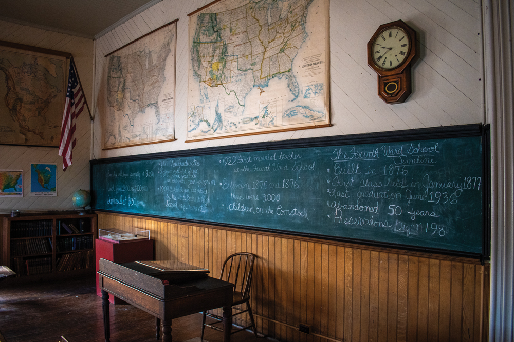 Green narrow chalkboard with handwritten cursive text about Fourth Ward School's history. There are two large maps above the chalkboard of the United States and there is a clock and a flag sitting next to them. On the wall adjacent to the chalkboard, there is another map of North America. In front of the chalkboard, there is a desk and a chair alongside a display case and a bookshelf with a globe on top of it. 