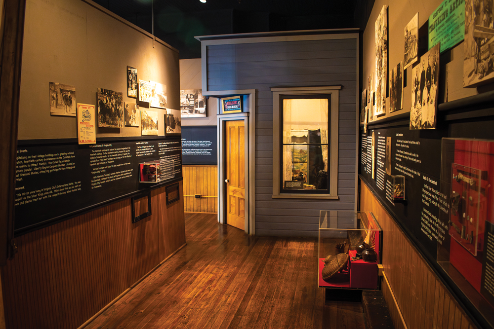 Museum Exhibit of Fourth Ward School. On the left hand side of the room, information about the school and pictures are displayed on the wall. Between the left hand wall and the back wall, there is a hallway that leads to another part of the the museum with more information, and a door sits on the corner of the back wall leading into another room with display cases. On the right hand wall, more pictures and information are displayed on the wall while a glass case sits pushed up to the wall, which features pottery. 