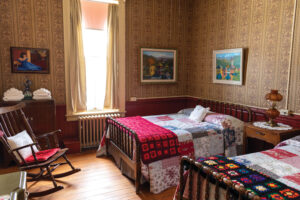 Photo of a bedroom in St. Mary's Art Center. There are two beds side by side and a night stand along the right hand wall, while a window, a dresser, and a rocking chair are along the left hand wall. The beds feature colorful quilts and a pillow. The nightstand has a lace doily with an antique lamp on top of it. There are three pictures on the walls, two on the left and one on the right. The first picture on the left is of a woman in a blue dress sitting on the ground. The second photo on the left is of a town with colorful, multistory buildings with mountains behind the buildings. The picture on the right is more colorful buildings with mountains behind. A adiator sits under the window, and the walls have a traditional floral wallpaper and a wooden wainscot chair rail along the bottom. On top of the dress are towels and a flower vase. The rocking chair has a red cushion pillow for sitting on and a white lace pillow for back support. 