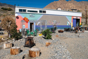 Exterior shot of Bizarre 101. A rectangular building with cinderblock walls sits as a background to a yard filled with rock, sand, and art pieces. The building is painted pink, purple, green, brown, blue, orange, and yellow. There is a metallic structure in front of a door on the left that hangs wind chimes. Underneath the metallic structure is a set of green outdoor furniture. There is foliage next to the metallic structure, and a stray reserved parking sign. In the lower left of the photo, there are four stumps sitting around a rectangular black fireplace that has wood inside it. On the right in front of the building, there are more outdoor funiture sets and other various indistinguishable items. An at piece sits in the middle of the building that features a wheel, a rams head, and bird like wings. Behind the building are mountains and another building. 