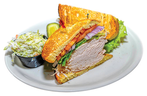 A plate with a piled-high turkey sandwich with lettuce, onions, and tomato. Pickle and coleslaw on the side.