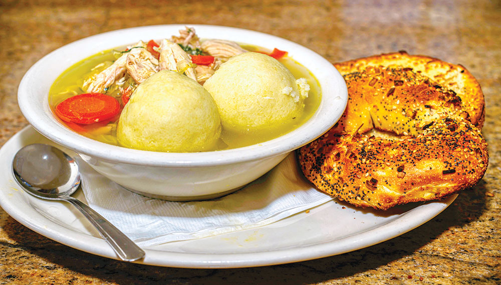 A plate with a bowl of matzoh ball chicken soup and an everything bagel beside it.