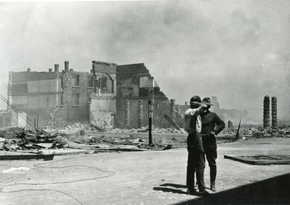 An old photo of two firemen standing on a street in front of a half ruined building with smoke rising from it. Photo was taken in 1923 Goldfield fire incident. 