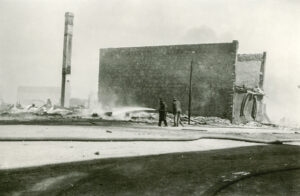 Two men can be seen with a firehose in their hands trying to put out a fire. Behind them stands a ruined building with brick walls still standing while everything else has been gutted on the building. 