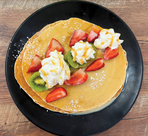 A stack of pancakes on a black plate and wooden table. Pancakes have strawberries, kiwi, whipped cream, caramel sauce, and powdered sugar on them.