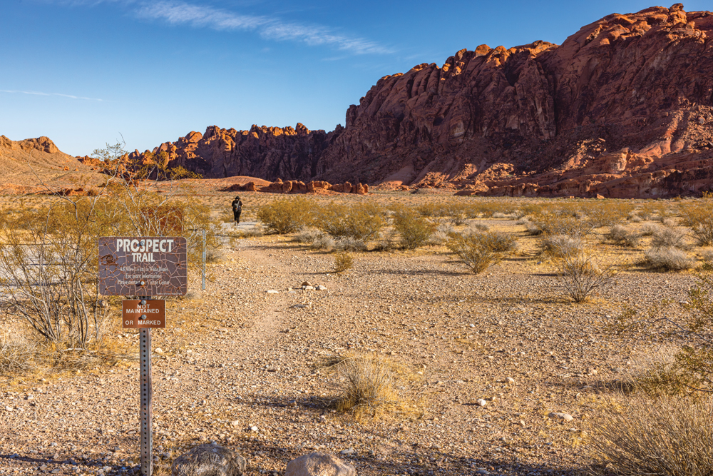 Woman walking towards towering red mountains along the Prospect Trail in Valley of Fire State Park. 
