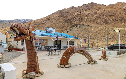 The front of Bighorn Crossing, with a metal sculpture of a dragon out front, and metal sculpture of two bighorn sheep fighting, off to the side. There are tables and chairs out front as well.