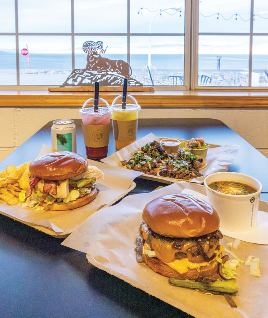 Burgers with chips, chili, tacos, and Italian sodas sitting on a table, with a metal cutout of a bighorn sheep on a rock in the window, and Walker Lake in the background, across the road.