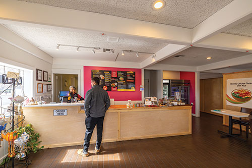 Inside of Pink Elephant restaurant, with ordering counter, menu, and hand-made teapot windchimes for sale. A customer is placing an order.