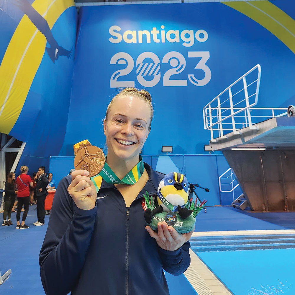 Olympian Krysta Palmer holding up a medal in front of a Santiago 2023 sign at a pool