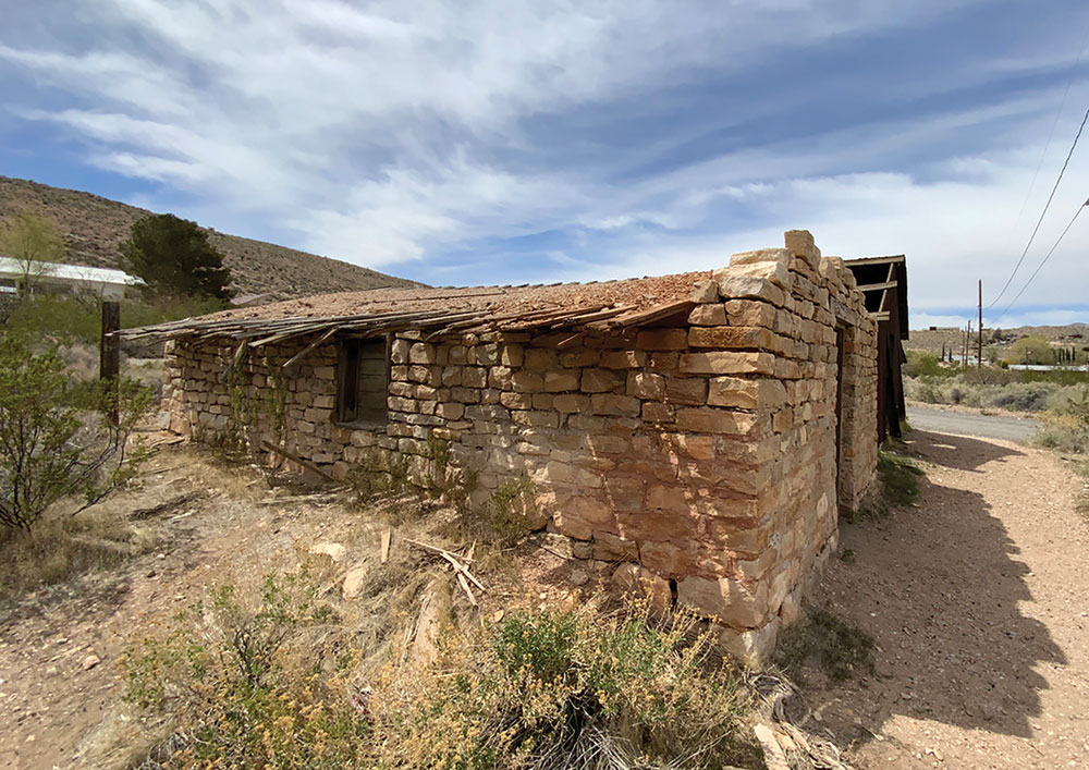 Photo shows the remains of a stone building, Yount Store, in the general stores site of the Goodsprings walking tour. There is sagebrush all around, and blue skies with clouds.