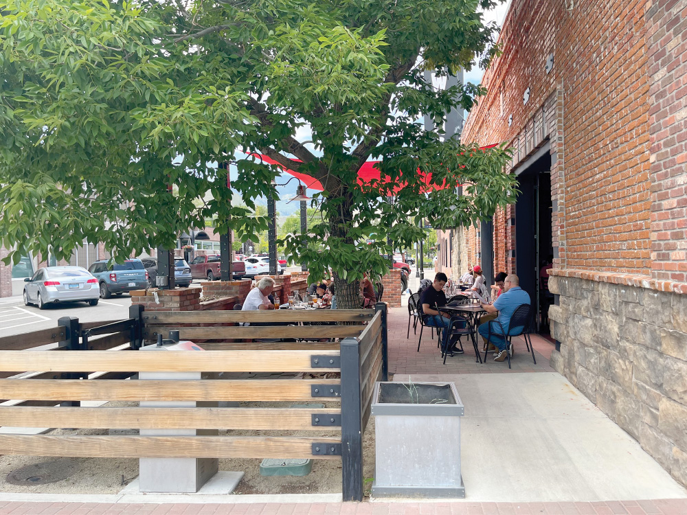 Exterior of Great Basin Brewing Company shows a brick building with a tree next to it, and many people seated outside, dining. Cars are parked nearby, across the street.
