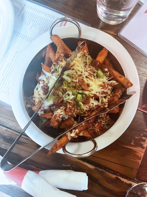 Sticky Fries at Great Basin Brewing Company. Fresh-cut sweet potatoes fried to perfection and smothered in BBQ sauce, white cheddar, bacon crumbles, and scallions, in a white bowl sitting on a distressed wooden table.