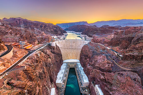 Hoover Dam at sunrise or sunset, with blue-green water, photographed from the longest viewing bridge in North America. Lots of red rock surrounds the large rock-gravity hydroelectric dam.